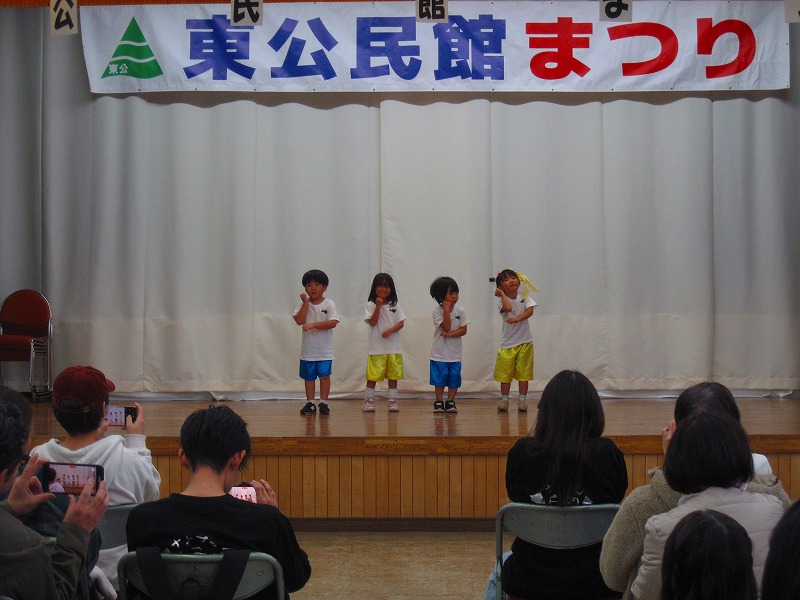 「東公民館まつり」と書かれた横断幕のある舞台の上で、4人の幼児が半袖シャツに半ズボンの衣装で立ち、手を頬にあてて考えるポーズを取っている写真