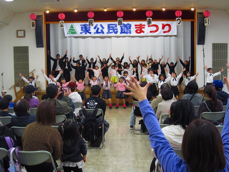 「東公民館まつり」と書かれた横断幕の舞台上で、様々な衣装を着た大勢の子供たちが両手を高く上げてダンスを披露している写真