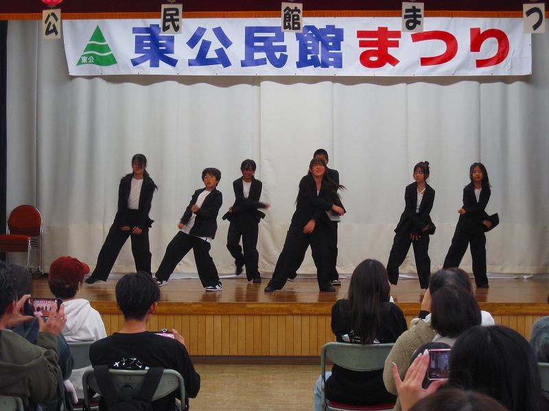 「東公民館まつり」と書かれた横断幕が掲げられた舞台の上で、白いシャツに黒い衣装を着用した7人組のグループが、激しい動きのダンスパフォーマンスを披露している写真