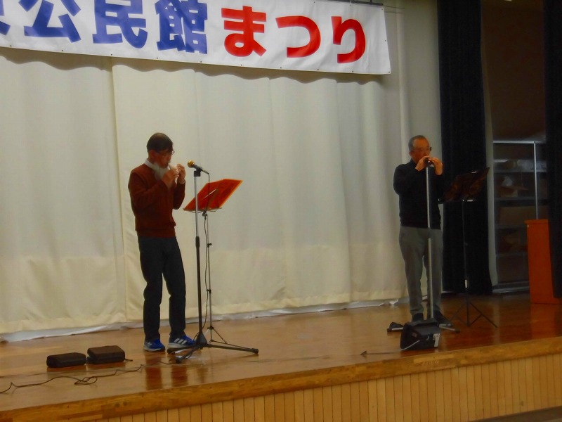 「公民館まつり」と書かれた横断幕の舞台の上で、2人の男性がそれぞれマイクスタンドの前に立ち、オカリナのようなものを吹いて演奏している様子を左側から写した写真