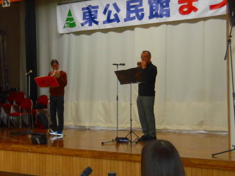 「公民館まつり」と書かれた横断幕の舞台の上で、2人の男性がそれぞれマイクスタンドの前に立ち、オカリナのようなものを演奏している様子を右側から写した写真