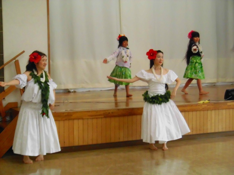 舞台に小学生くらいの女の子2人と、舞台前には2人の女性が赤い花飾りを付けてフラダンスを披露している写真