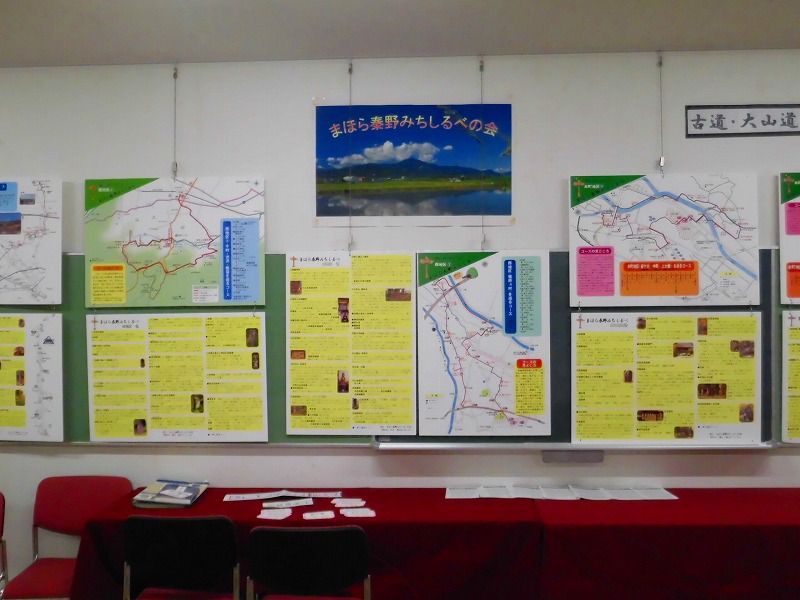 壁に「まほろば秦野みちしるべの会」と書かれたポスターを中心に、地域の地図や歴史、観光に関する情報が記載された複数のパネルが展示されており、手前の赤いクロスが敷かれた机には資料が置かれている展示の様子の写真