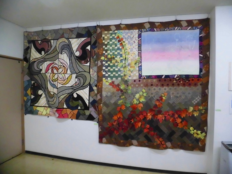白い壁に2枚の大きなパッチワークキルト作品が展示されており、左の作品はステンドグラスのような幾何学模様、右の作品は夕焼け空と紅葉したツタが描かれた風景画をモチーフにした写真