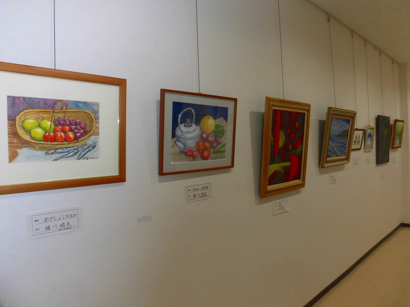 白い壁に、かごに盛られた果物の絵画などが横一列に吊り下げて展示されており、絵の下には作者名と作品名が書かれたキャプションが貼られている展示会場の様子の写真