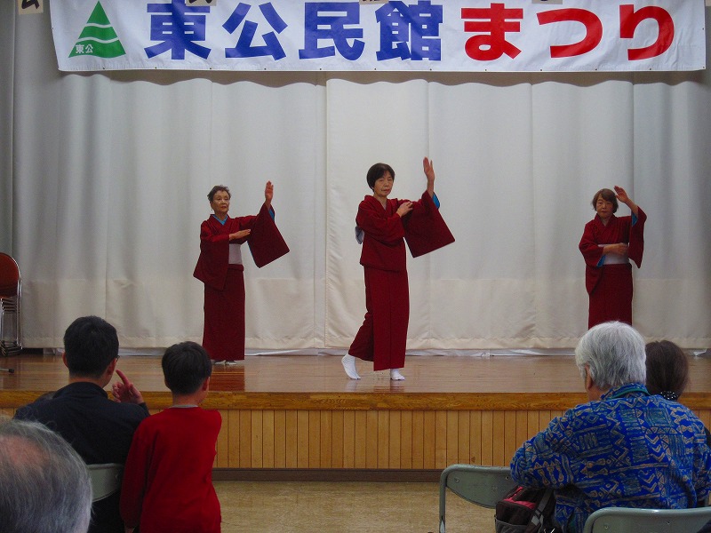「東公民館まつり」と書かれた横断幕の舞台の上で、赤い着物を着たの3人の女性が日本の伝統舞踊らしき優雅な踊りを披露している写真