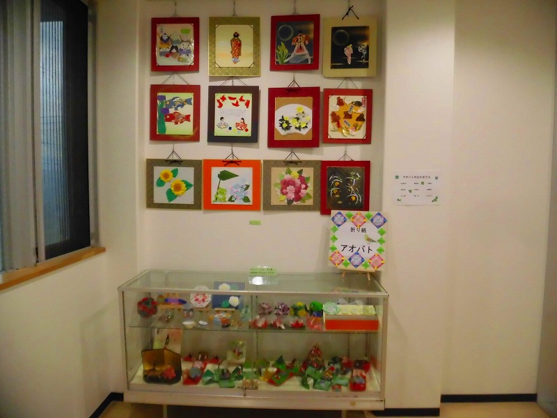 季節をテーマにした貼り絵が壁に展示されており、ガラスケースには様々な色の折り紙作品が並べられている写真