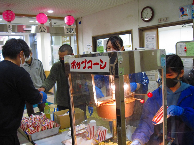 青い手袋を着用したスタッフ数名がポップコーンの機械からポップコーンを袋に詰めており、お客さんに手渡している様子の写真