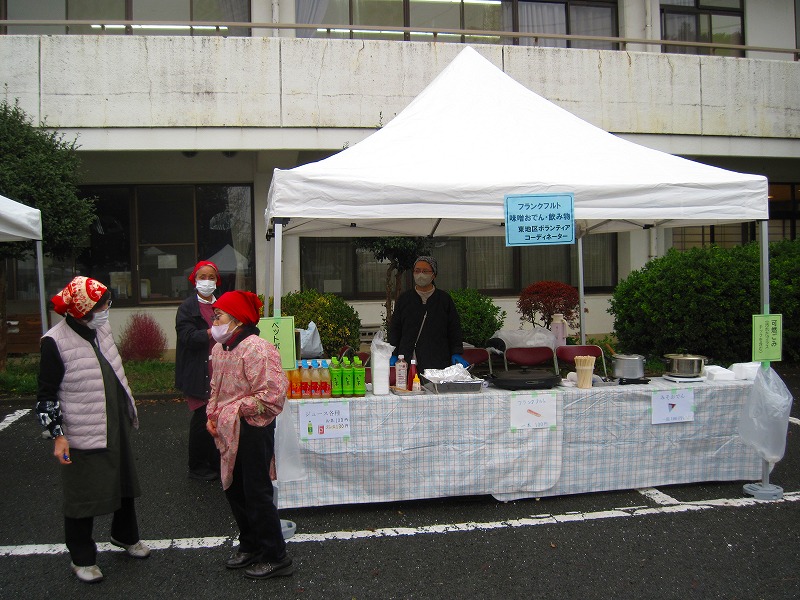 野外の白いテントの下で、机の上にお茶やジュースなどが並べられており、赤いバンダナとマスクを着用した女性たちがブース前で話をしている写真