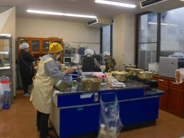 三角巾やエプロンを着用した数名の女性たちが、調理台の上で複数の大きな寸胴鍋を使い、料理の仕込みや準備をしている写真