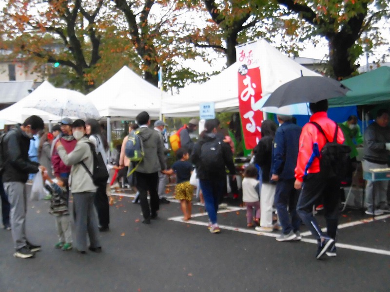 小雨の降る曇天の下、白いテントにさまざまなブースが並んでおり、多くの人々が飲食や買い物を楽しんでいる写真