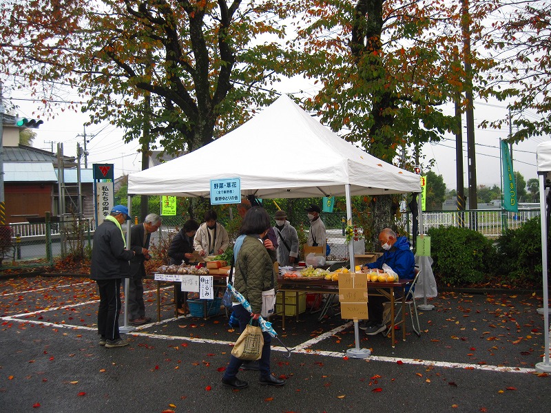 屋外の駐車場に白いテントが設置され、野菜や草花などを販売するブースがあり、来場者が商品を見たり購入したりしている様子の写真