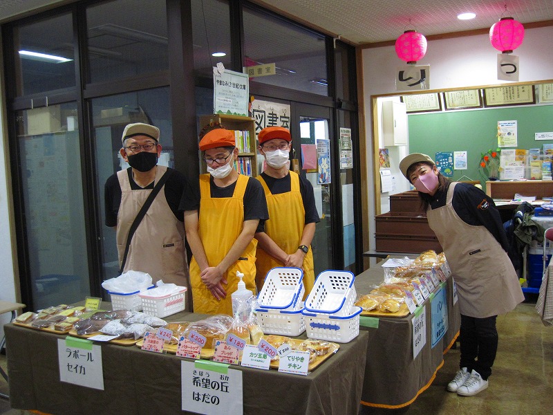 エプロンを着用した4人のスタッフが、パンや焼き菓子などを並べた販売ブースのカウンターに立ち、笑顔で接客している様子の写真