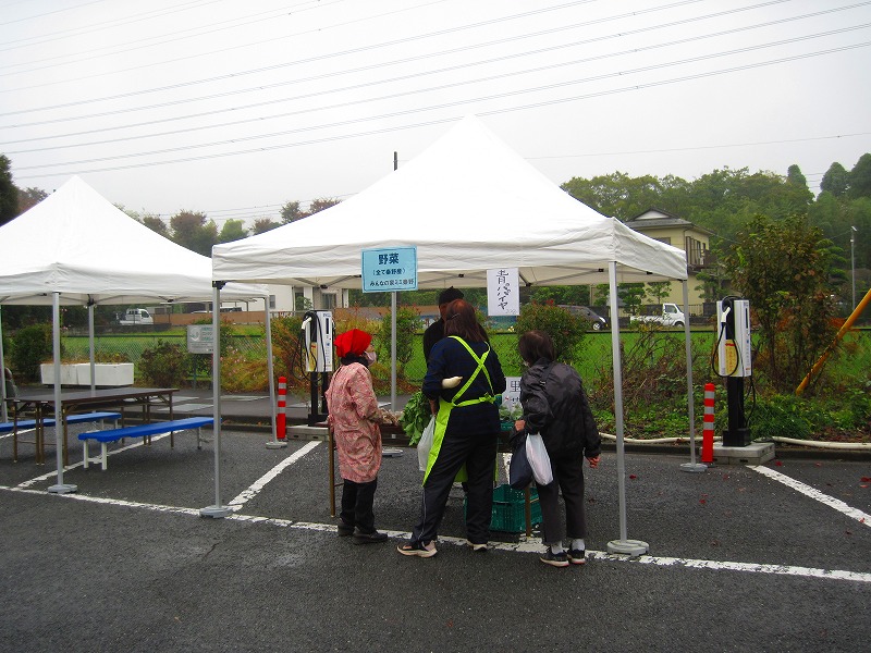 曇り空の下、駐車場に設置された白いテントの下で、野菜を販売するブースにお客さんが集まっている写真