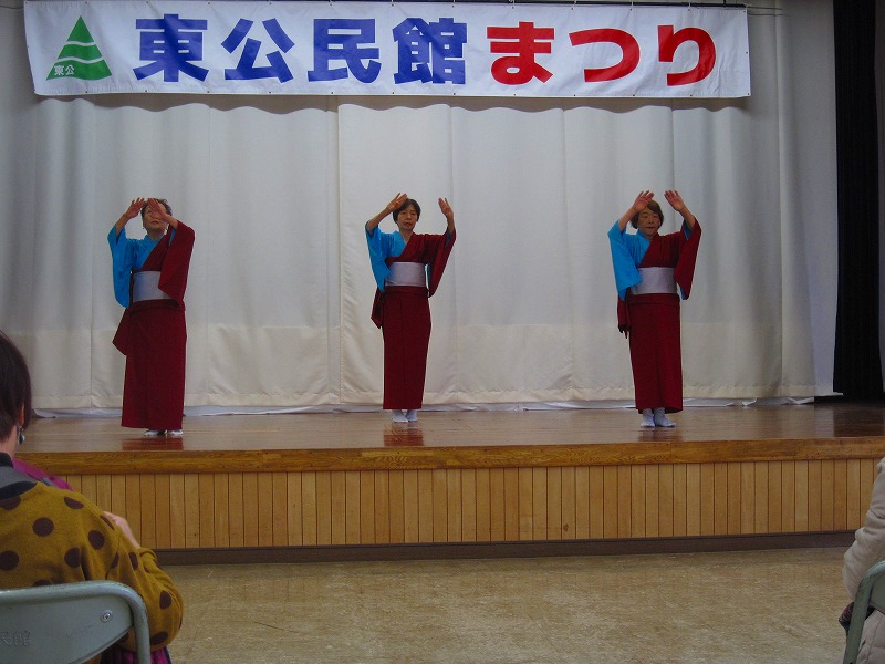 「東公民館まつり」と書かれた横断幕の舞台の上で、赤い着物を着た3人の女性が手を前に出し、伝統舞踊らしき優雅な踊りを披露している写真