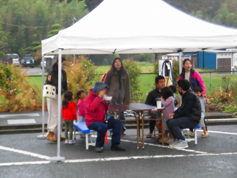 駐車場に立てられた白いテントの下で、男性や女性、子供達が椅子に座り、飲食を楽しんでいる様子の写真