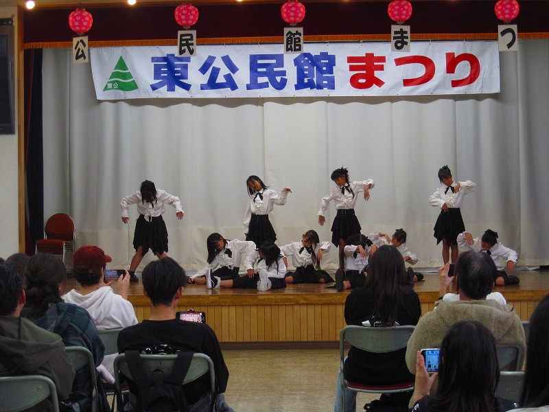 「東公民館まつり」と書かれた横断幕がある舞台の上で、白と黒のお揃いの衣装を着用した10人ほどの子供たちが、ポーズを取りダンスを披露している写真