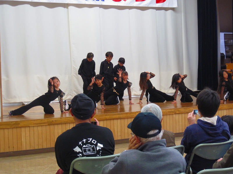 舞台の上で、黒の衣装を着用した小学生くらいの子供達が、足を延ばしたり手を広げたり、躍動感あるダンスをしている写真