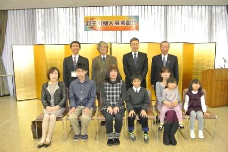 「親子川柳大会表彰式」の横断幕が掲げられた窓の前に設置した金屏風の前で、前列の椅子に3組の親子が座り、後列にスーツ姿の男性4人が並んで立って写した記念写真