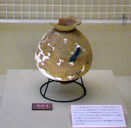 底に三脚の展示台を置き、ひび割れ部分を白い漆喰のようなもので修復した茶色い土器が展示されている様子の写真
