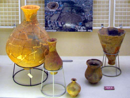 再構成された大小様々な土器が展示台に並べられ、その背後には発掘現場を示す航空写真が掲示されている写真