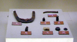 ガラスケース内に、錆びた鉄製の農具や小さな石の玉、不明な遺物など、複数出土品が並んで展示されている様子の写真