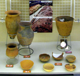 茶褐色の土器と土器片、および出土状況を解説する掲示物が、展示ケースに整然と並べられている様子の写真