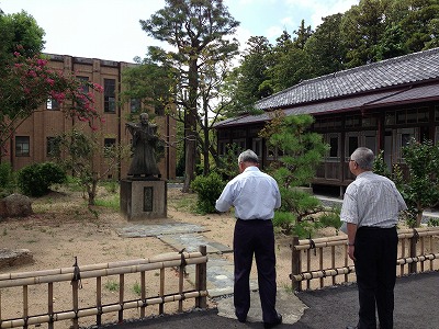 右側に平屋、奥に2階建ての建物がある手前の中庭に、甲冑姿の武士の像が立っており、2人の男性が竹の柵越しに像を眺めている写真