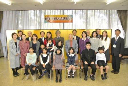 「親子川柳大会表彰式」の横断幕が掲げられた窓の前に設置した金屏風の前で、前列の椅子に6人の子どもたちが座り、その後ろにたくさんの出席者が並んで立って写した記念写真