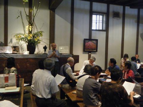 たくさんの人々が机に向かい、資料を見ながら、大きな生け花が飾られた壁の前に立つ男性の説明を聞いている様子の写真