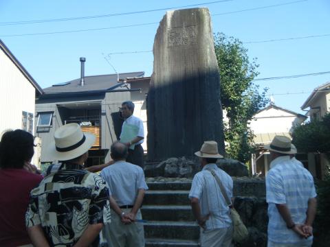 複数の参加者たちが、巨大な石碑の周りに集まり、階段の上に立った講師の男性の説明を聞いている写真