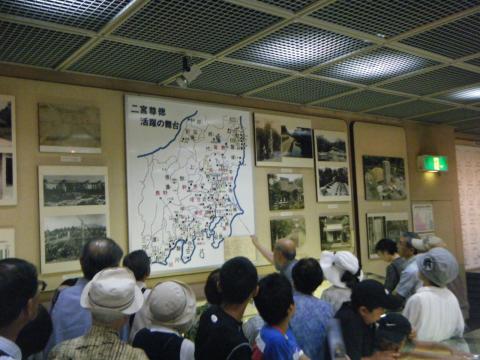 博物館の壁に大きな地図とたくさんの写真パネルが展示されており、館長代理の男性が棒で地図を差しながら、集まった参加者たちへ説明をしている様子の写真