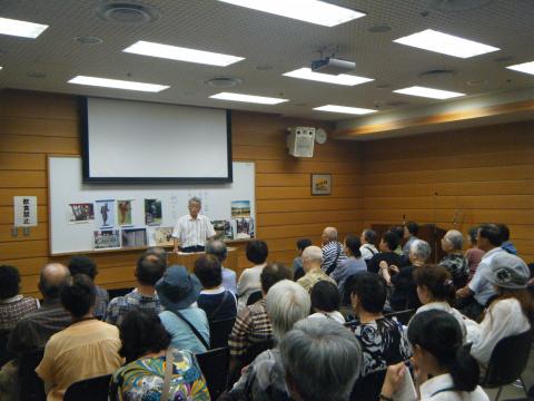 多数の参加者が教室で机に向かって座っており、前方で講師の男性が、地図が貼られた黒板を指さしながら話をしている写真