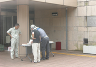 大きな建物の入り口前に設置したテーブルを3人の男性が囲んでいる様子の写真