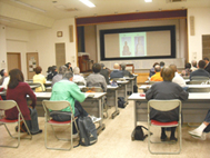 広い室内で多数の受講者が椅子に座って前方にある大きなスクリーンを見つめており、講義が行われている様子を捉えた写真