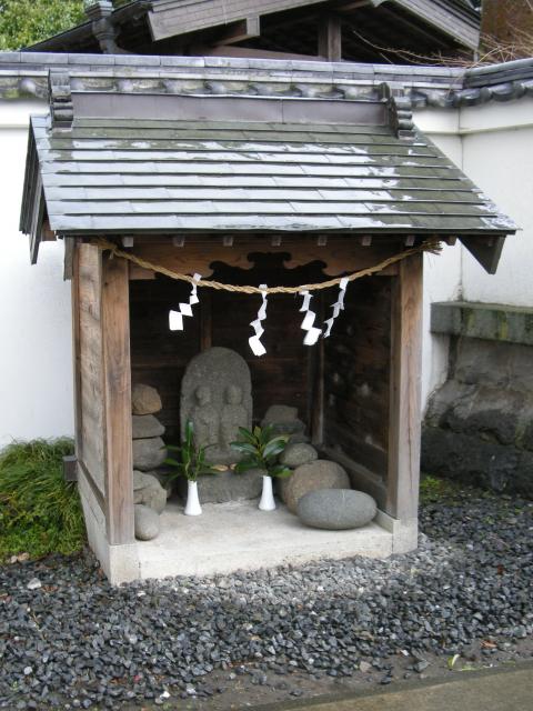 白壁の前の砂利敷きの一角にある小さな祠の中に、双体道祖神や複数の丸い石が安置されている、戸川原の双体道祖神の写真