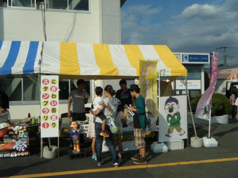 建物前に、「にぎわい街巡りスタンプラリー」と書かれた立て看板が設置された、青と白、黄色と白のストライプ模様の屋根のテントが並び、親子連れの来場者がテントの中の係りの男性たちから説明を受けている様子の写真