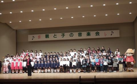 「親と子の音楽会」と書かれた横断幕が掲げられた舞台上で、大勢の大人と子どもが合唱隊として並び、客席を向いて指揮者が立っている写真