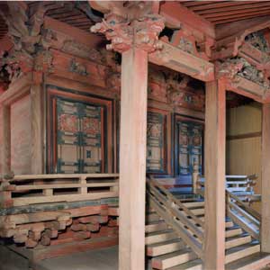 塗りの柱が垂直に立ち、複雑な木組みが支える屋根の下に、木製の手すりがついた階段があり、その奥に装飾が施された古い扉が見える八幡神社本殿の写真