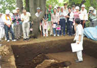 四角く掘られた深い穴の前に多数の見学者が集まり、穴の中に立った男性が説明を行っている、発掘調査現地見学会の様子の写真