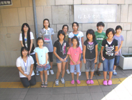 参加者の子どもたちが、屋外の白い壁を背景に外でカメラに向かって並んで立っており、左端に一人の女性がしゃがんで写っている集合写真