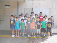 たくさんの参加者の子どもたちが、屋外の白い壁を背景に並んでカメラに向かって立っている集合写真