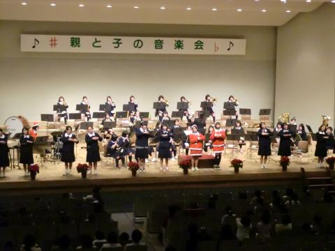 「親と子の音楽会」と書かれた横断幕の下、、黒の制服や赤いサンタ衣装を着た数十人の小中学生が合奏と合唱を披露し、多くの観客が着席している写真