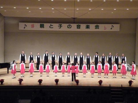 「親と子の音楽会」と書かれた横断幕の下、白いシャツと黒いベストを着た大人と、ピンクの衣装を着た子供や女性がステージ上で合唱する様子を捉えた写真