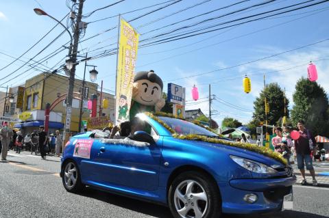 人々で賑わう、提灯が飾られた祭り会場前の通りを、黄色いのぼりを持った「「にのみやそんとくん」のマスコットを乗せた青いオープンカーが走っている様子の写真