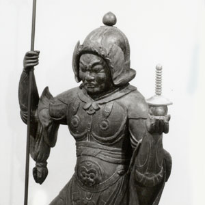 兜と鎧を身に着け、右手に長い棒、左手に宝塔を持った墨色の木造毘沙門天立像の写真