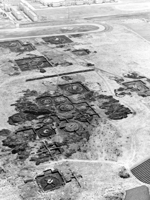 広い土地に円形または方形の区画が多数確認できる、大規模な遺跡の発掘現場を俯瞰で撮影したモノクロの航空写真