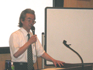 ネクタイを着用した山口先生がマイクを持って壇上で発表を行っており、その後ろには白いスクリーンが配置されている、講座風景を撮影した写真