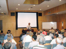 ホワイトボード横に設置されている大きなスクリーンの前に立った山口先生が、席に座った大勢の受講者に向かって話をしている、講座の様子の写真