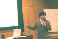 ホワイトボードの前立った加部先生がマイクを持ち、後方の大きなスクリーンに手を向けながら受講者の方を向いて話をしている写真